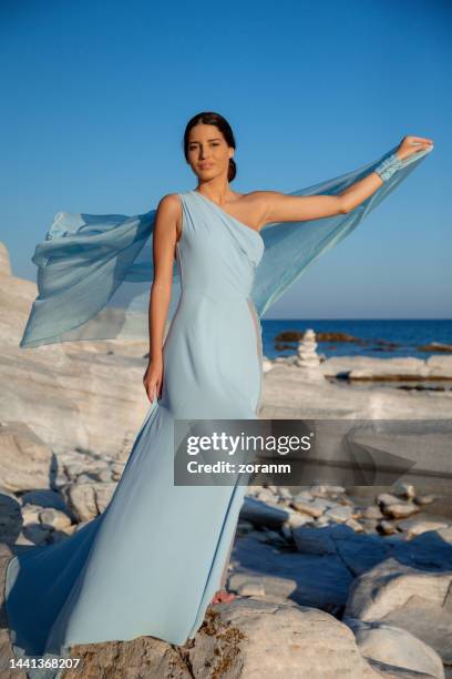 beautiful young woman in long light blue sleeveless dress playing with silky gown on the rocks by the sea - women in long dress stock pictures, royalty-free photos & images