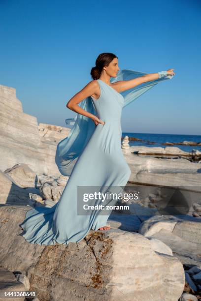 beautiful young woman in long light blue sleeveless dress playing with silky train on the rocky coastline - woman long dress beach stock pictures, royalty-free photos & images