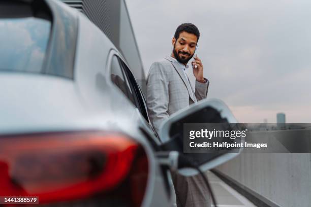 young multiracial man waiting for car charging. - elektroauto tanken stockfoto's en -beelden