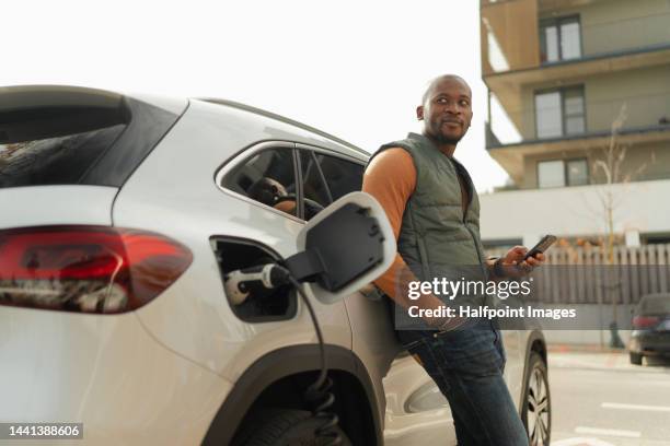 young multiracial man waiting for charging his car. - hybrid car charging stock pictures, royalty-free photos & images