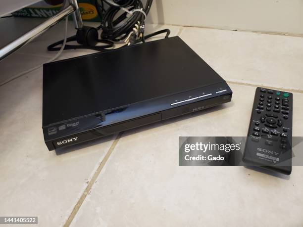 High-angle shot of a black Sony DVD player and remote, with Sony, Dolby, and HDMI logos, resting on a white tiled floor with cords and a media cart...