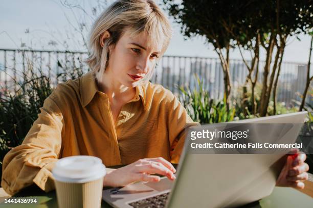 a woman sits outside on a balcony and works on her laptop, a cup of coffee sits beside her. - author laptop stock pictures, royalty-free photos & images