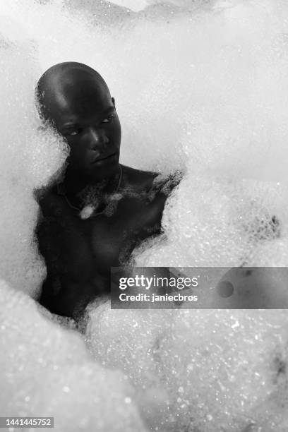handsome african ethnicity muscular man relaxing in the bath covered by foam. self-care and wellness metaphor. vertical image grayscale - monochrome bathroom stock pictures, royalty-free photos & images