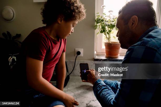 father showing smart phone screen to son sitting on kitchen counter at home - child on phone stock pictures, royalty-free photos & images