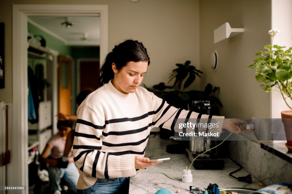 Woman charging smart phone while standing near kitchen counter at home
