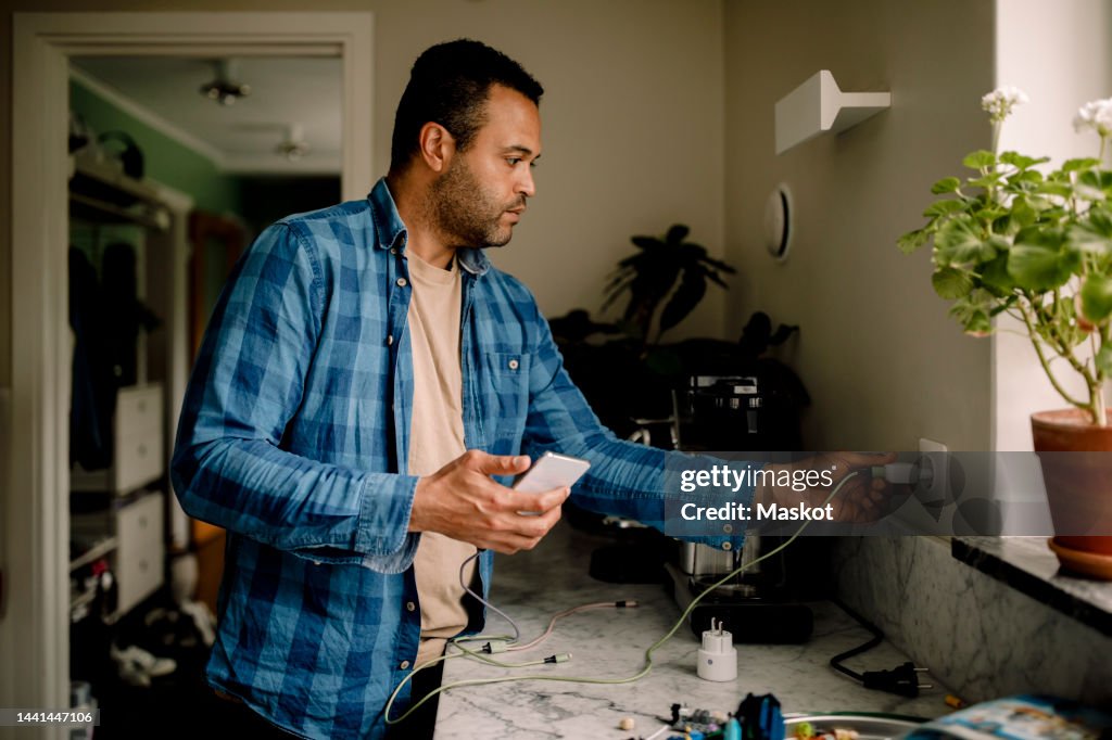 Man plugging smart phone charger in power socket while standing near kitchen counter at home