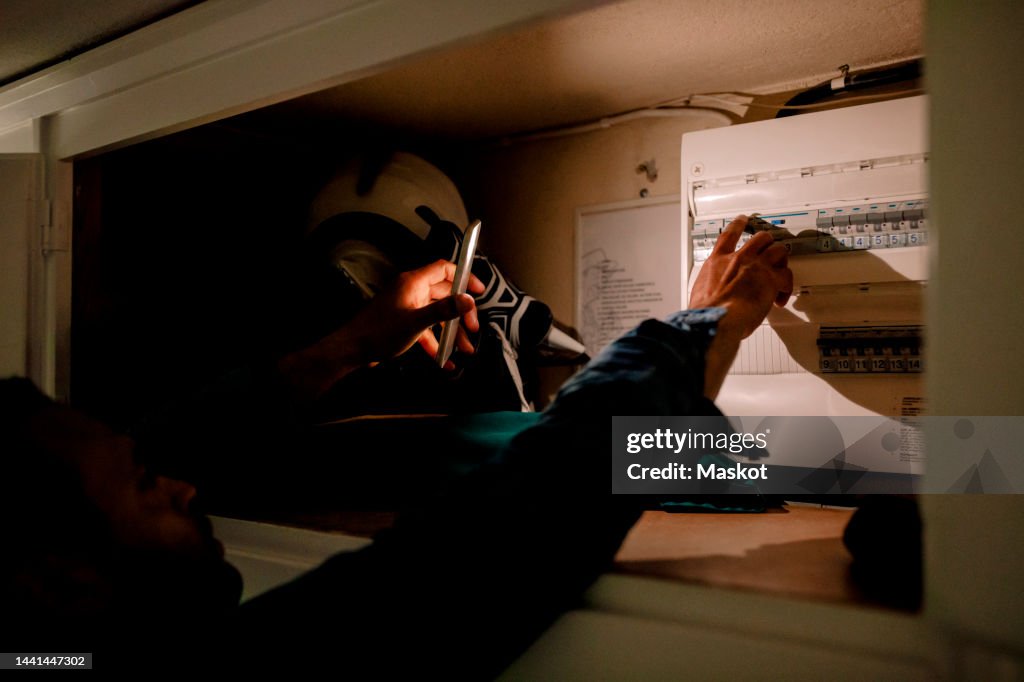 Man turning on switch from fuse box using smart phone torch during blackout