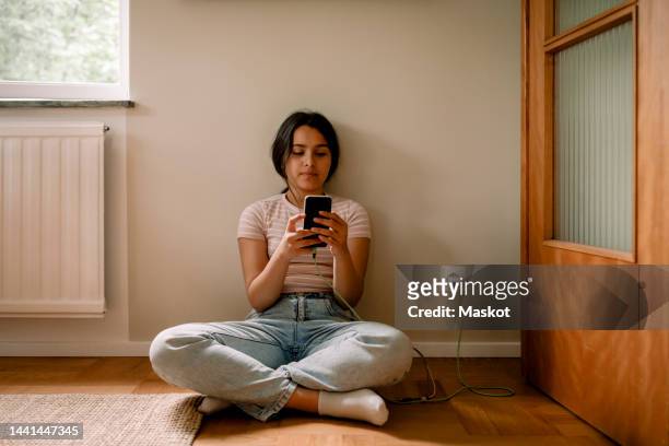 girl using smart phone while sitting cross-legged on floor against wall at home - social media addiction stock pictures, royalty-free photos & images