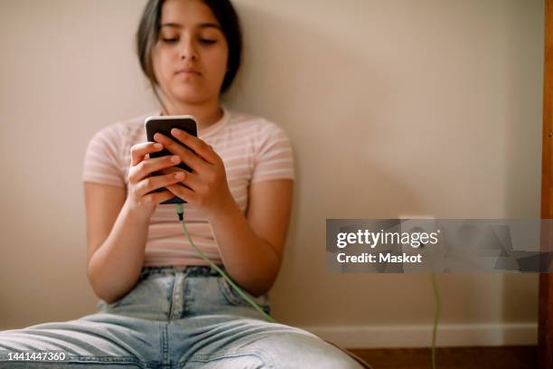 girl charging and using mobile phone while sitting against wall at home - chargeur de batterie photos et images de collection