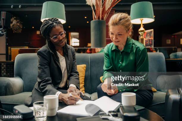 young businesswoman talking with female colleague while signing contract in hotel lounge - businesswoman hotel stock pictures, royalty-free photos & images