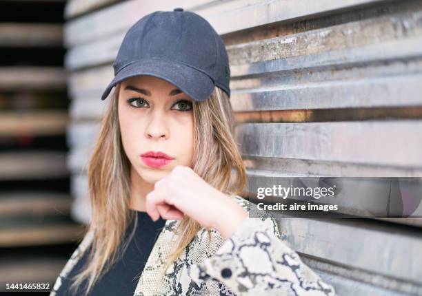 portrait of a young woman wearing a baseball cap and a leopard blazer on a sheet metal background - metallic blazer stock pictures, royalty-free photos & images