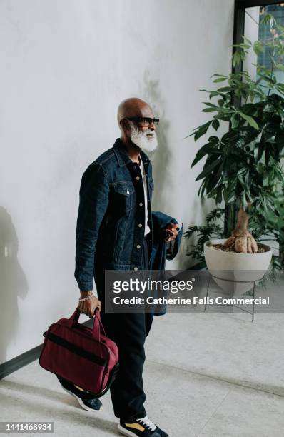 a stylish, confident older gentleman holds a bag and walks through a modern interior space - mature men walking stock pictures, royalty-free photos & images