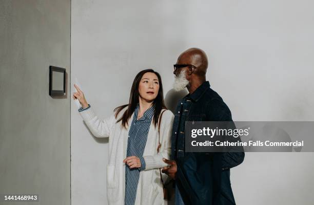 a woman explains a wall mounted electronic device to a man, who listens intently. - elektrogeschäft stock-fotos und bilder