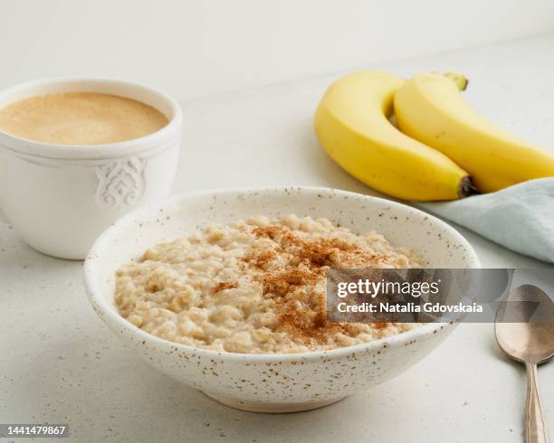 oatmeal porridge with cinnamon, cup of coffee cappuccino, bananas. traditional bunting breakfast served in ceramic bowl on gray background. vegetarian and nutritious vitamin morning eating. classic hot homemade dietary meal - harina-de-avena fotografías e imágenes de stock
