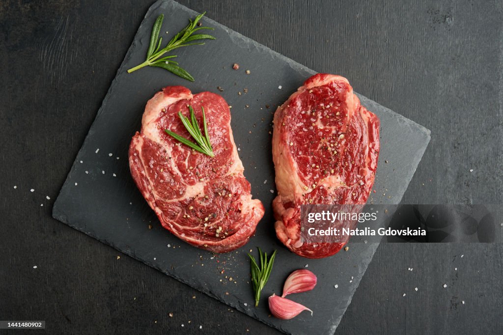 Ribeye, rib eye steak, raw beef meat tender marbled striploin steak on dark background, with salt, garlic clove and spices rosemary on black stone board. Uncooked sirloin. Preparation for cooking