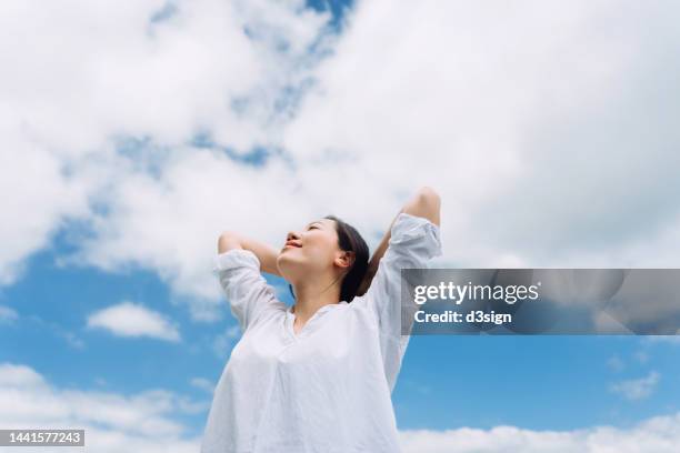 low angle portrait of relaxed young asian woman with her eyes closed stretching arms in the nature, setting herself free and feeling relieved. enjoying fresh air and sunlight with head up against blue sky. freedom in nature. connection with nature - arms raised to sky stock pictures, royalty-free photos & images