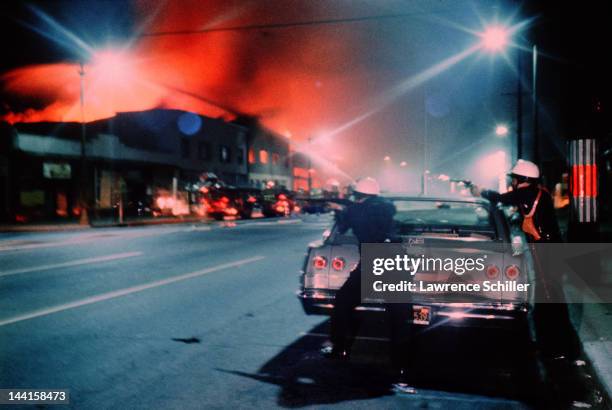 During the Watts Riots, two police officers take aim from behind a parked car as fire and smoke from a burning building color the night sky, Los...