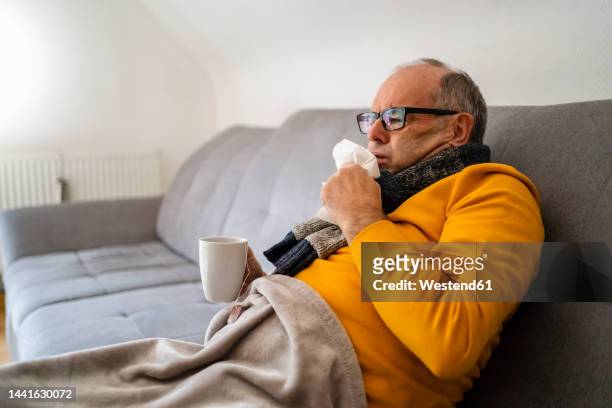man coughing sitting on sofa in living room - influenza virus stock pictures, royalty-free photos & images