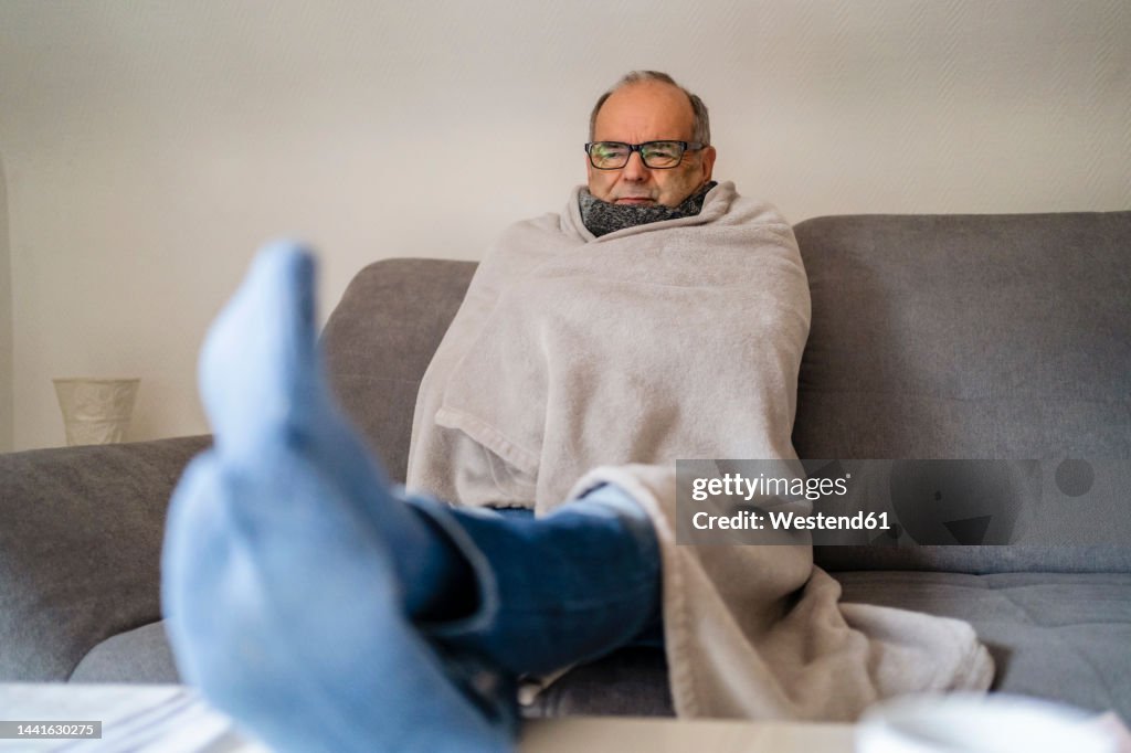 Senior man wrapped in blanket sitting on sofa at home