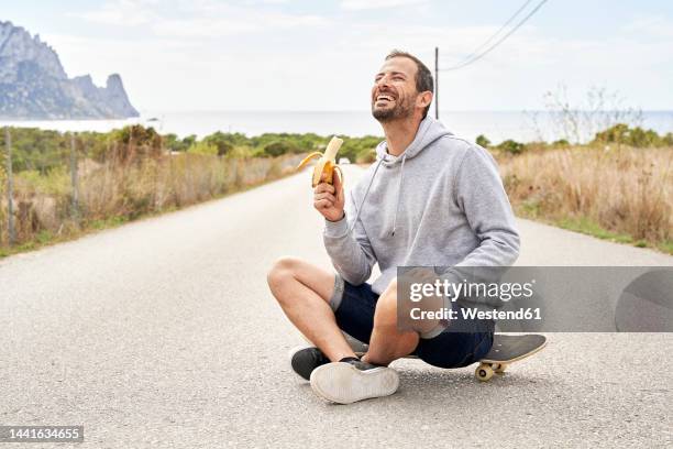 cheerful man with banana sitting on skateboard - banane essen stock-fotos und bilder