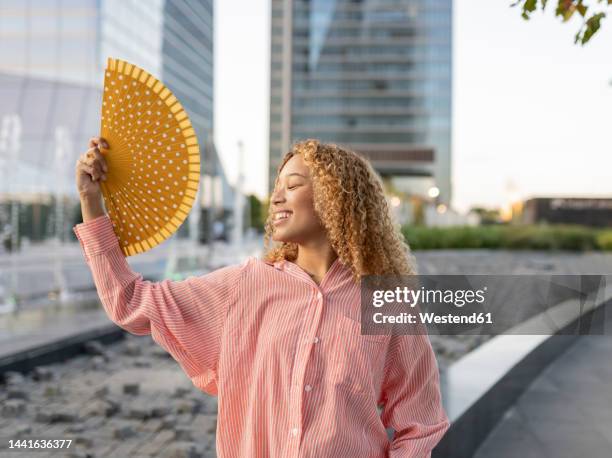 smiling woman with eyes closed using folding fan on street - waaier stockfoto's en -beelden