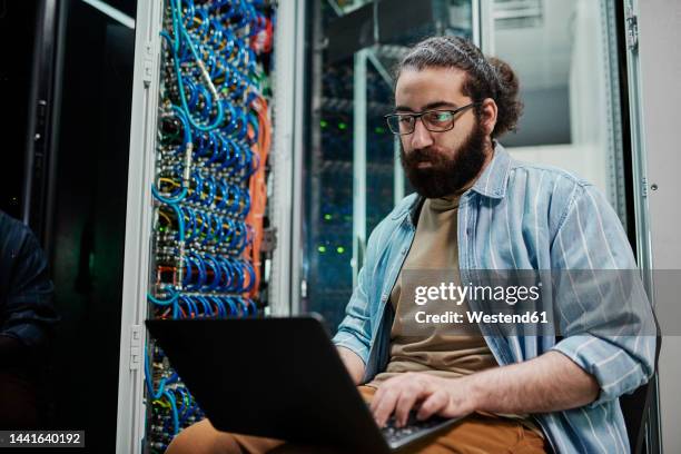 it expert working on laptop sitting in server room - server stock-fotos und bilder