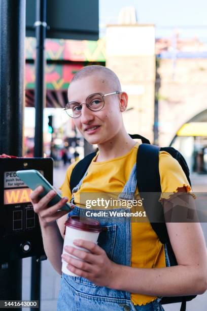 non-binary person with eyeglasses holding disposable coffee and mobile phone - androgynous stock-fotos und bilder