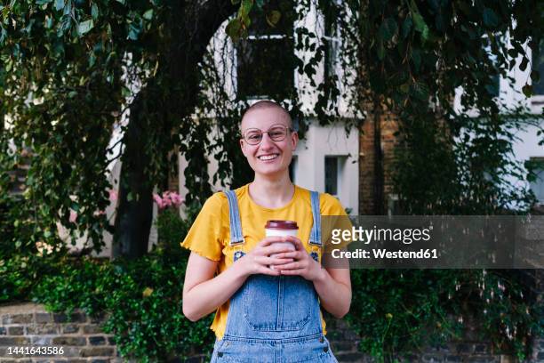 happy non-binary person with disposable coffee cup in front of plants - androgyn stock pictures, royalty-free photos & images
