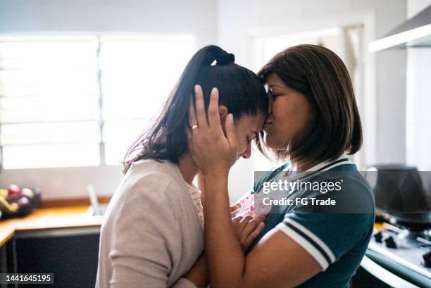 mid adult woman kissing the forehead of the her wife in the kitchen at home - images of lesbians kissing stock pictures, royalty-free photos & images