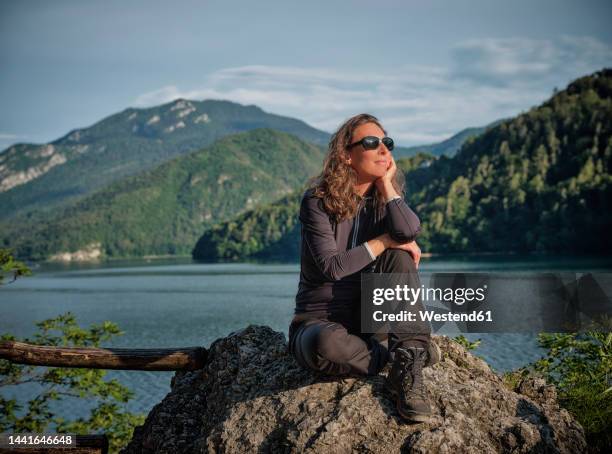 smiling thoughtful woman sitting on rock in front of lake - trentino stock pictures, royalty-free photos & images