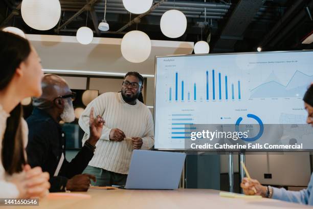 a young male gives a presentation, casting onto a large monitor from his laptop. - business presentation stock pictures, royalty-free photos & images