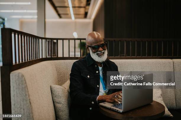 a older gentleman sits in a booth on his laptop - black businessman stock pictures, royalty-free photos & images