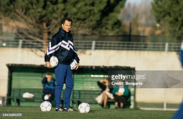 German football manager and former footballer Franz Beckenbauer, holding two footballs, one under each arm his hands in his pockets, wearing a...