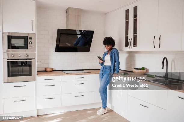 young woman using smart phone leaning on kitchen counter at home - leaning stock pictures, royalty-free photos & images