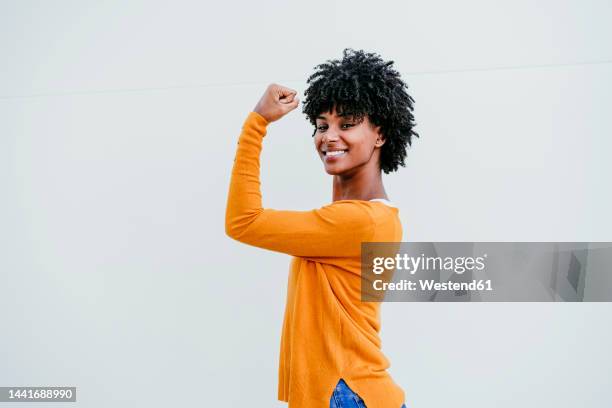 smiling young woman flexing muscle near white wall - flexing muscles stock pictures, royalty-free photos & images