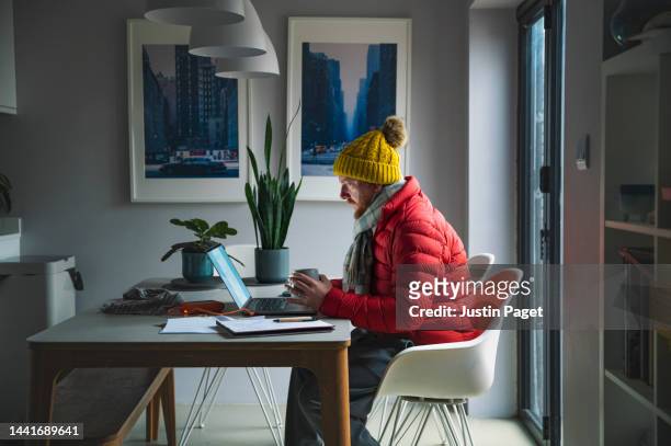 mature man sitting working from home in a red puffer coat, scarf and wooly hat - cost of living crisis - froid photos et images de collection