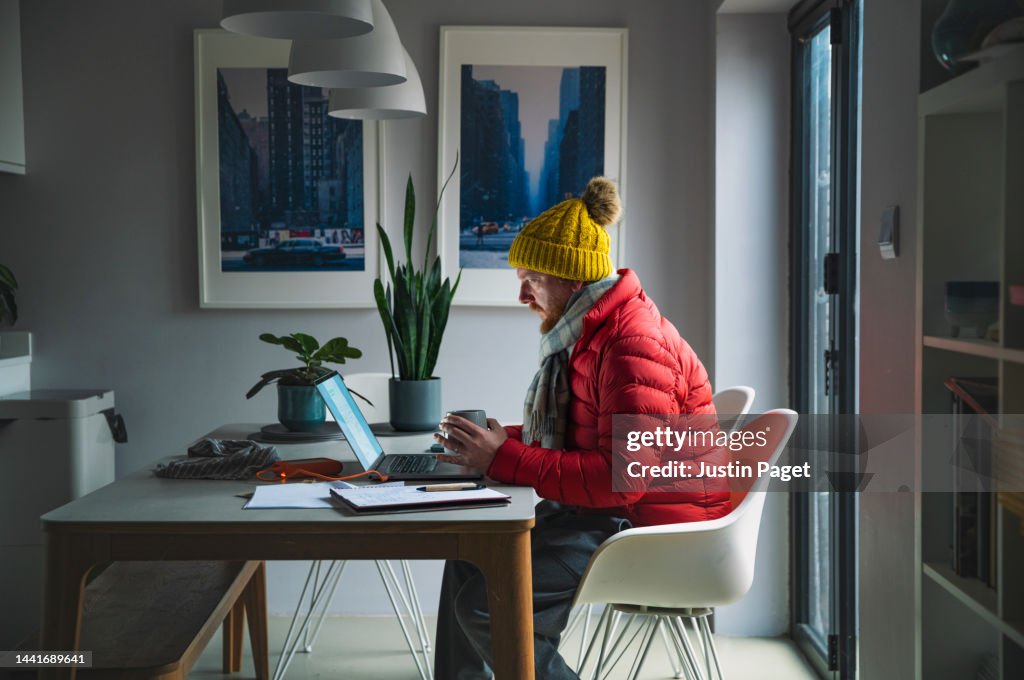 Mature man sitting working from home in a red puffer coat, scarf and wooly hat - Cost of Living Crisis