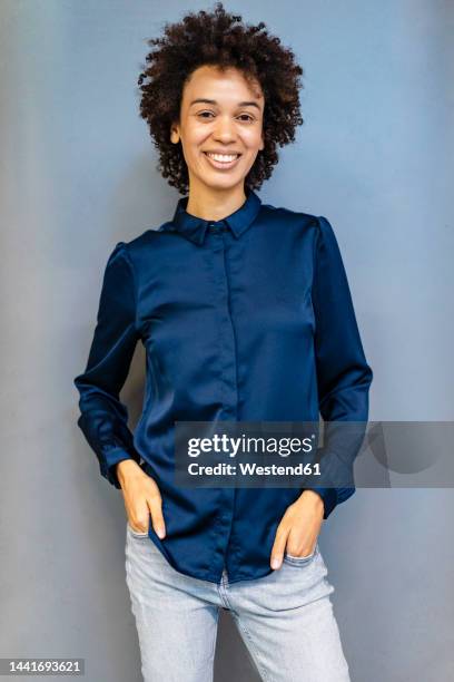 smiling businesswoman with hands in pockets standing against blue background - bluse stock-fotos und bilder