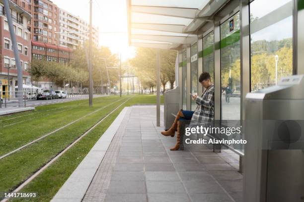 woman sitting waiting for public transport in a city. care for the environment - bushaltestelle stock-fotos und bilder