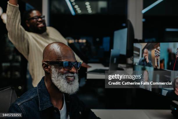 two men work late at the office. one takes a video conference call, while the other comically interrupts the meeting to wave at the people on the screen. - boss over shoulder stock pictures, royalty-free photos & images