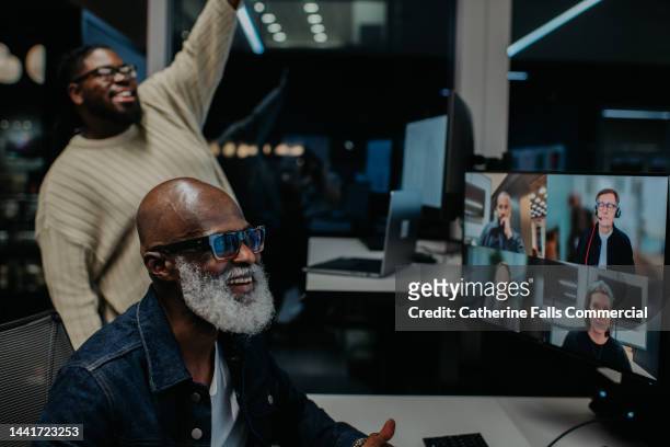 two men work late at the office. one takes a video conference call, while the other comically interrupts the meeting to wave at the people on the screen. - boss over shoulder stock-fotos und bilder