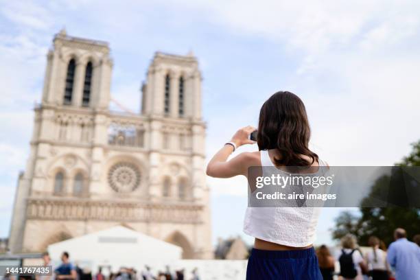 girl in casual clothes standing taking pictures towards the cathedral of notre dame on a sunny day, low angle back view - church color light paris stockfoto's en -beelden