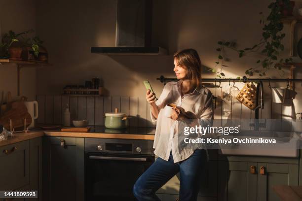 young woman using smartphone leaning at kitchen table with coffee mug and organizer in a modern home. smiling woman reading phone message. brunette happy girl typing a text message - smart kitchen fotografías e imágenes de stock