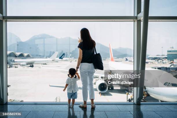 rear view of young asian mother holding hands of her cute little daughter, looking at airplane through window at the airport terminal while waiting for departure. family travel and vacation. embark on a new journey with the family - tochter zeigt stock-fotos und bilder