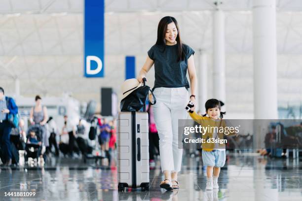 joyful young asian mother holding hands of her cute little daughter, walking through airport concourse and travelling by plane on a vacation. family travel and vacation. embark on a new journey with the family - family at airport fotografías e imágenes de stock