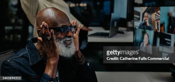 a man on an online video call touches the sides of his glasses and smiles - person surrounded by computer screens stock-fotos und bilder