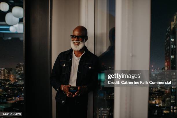 a man stands against a window at nighttime holding a phone, he glances up at the camera and laughs. - tinted eyeglasses stockfoto's en -beelden