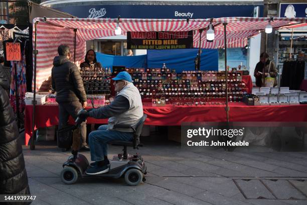 An elderly man rides a mobility scooter past a stall selling watches in Barking Town Centre street market on November 14, 2022 in Barking, London.