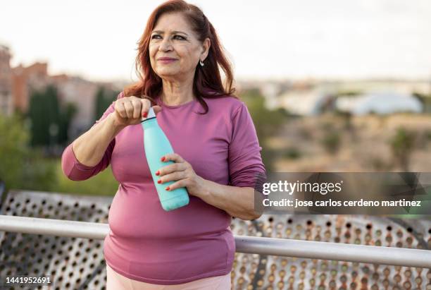 senior plus size hispanic woman drinking a bottle of water in a public park - plus size women stock-fotos und bilder