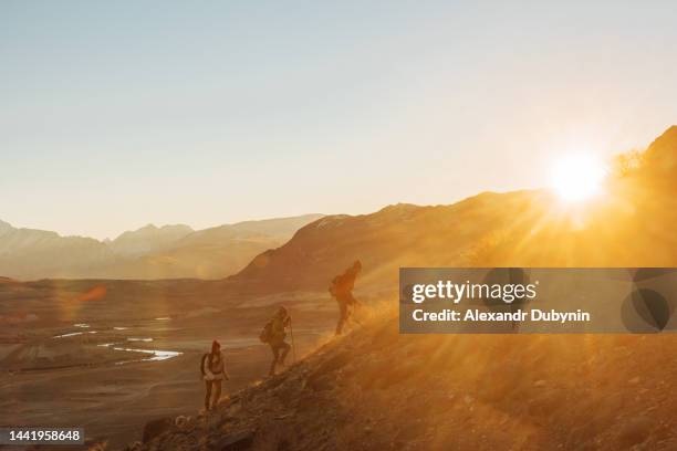 a group of hikers with backpacks walking uphill at sunset to the top of the mountain. travel and adventure concept - discovery bags walking stock-fotos und bilder